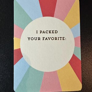 K&ouml;nnte beinhalten: Eine farbenfrohe Karte mit einem radialen Design in Rot, Rosa, Gelb, Blau und Gr&uuml;n. Die Karte hat einen wei&szlig;en Kreis in der Mitte mit dem Text "I PACKED YOUR FAVORITE:" in Schwarz gedruckt.