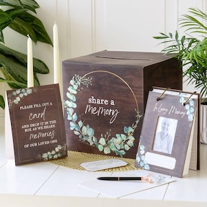 May include: A dark wood-grain memory box with a gold circular eucalyptus wreath and the text share a memory. Includes a matching sign reading please fill out a card and drop it in the box as we share memories of our loved one, and a photo frame.
