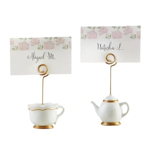 May include: Two place card holders with white cards featuring names and floral accents. One holder is a white teacup with gold trim, the other a white teapot with gold trim. Both have gold wire card holders.