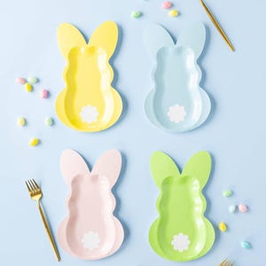 May include: Four pastel-colored bunny-shaped paper plates in yellow, blue, pink, and green, each with a white flower-shaped tail. Gold forks and colorful candies are scattered on a light blue background. Easter-themed tableware.