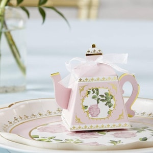 May include: A pink, decorative teapot-shaped box with gold trim and a white ribbon. The box features a floral design with a pink rose and green leaves. It sits on a floral-patterned plate.