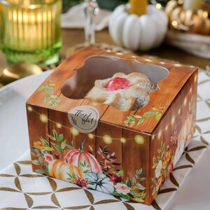 May include: A square cardboard bakery box with a clear window, featuring a rustic wood-grain print, autumn pumpkin illustrations, floral accents, and a string of lights. A round sticker with the text Thank you is attached to the top lid.