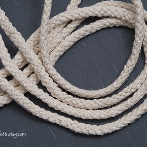 May include: Close-up of natural white cotton rope, braided in a thick, textured pattern. The rope is coiled on a dark grey surface.