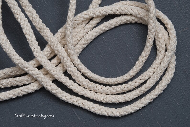 May include: Close-up of natural white cotton rope, braided in a thick, textured pattern. The rope is coiled on a dark grey surface.