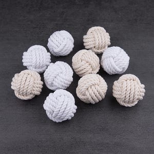 May include: A collection of ten spherical rope knots arranged in a cluster. The knots are crafted from twisted cord in alternating shades of bright white and off-white cream, each featuring a complex, interwoven monkey fist knot pattern.