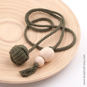 以下が含まれることがあります： オリーブグリーンのロープネックレス。結び目のボール、木製のビーズ、タッセルが付いています。ネックレスは、明るい茶色の木製プレートに置かれています。シンプルで自然なデザインです。