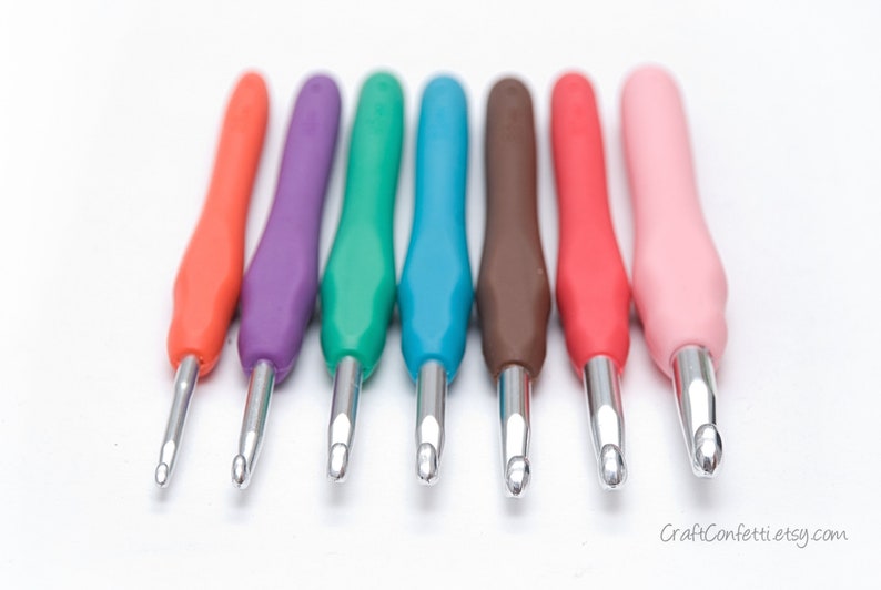 May include: A set of seven colorful crochet hooks with silver tips. The hooks are arranged in a row from left to right, with the orange hook on the left and the pink hook on the right.