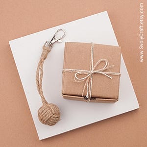 May include: A small, square, brown cardboard gift box tied with twine, next to a keychain with a rope ball. The keychain has a silver clasp. The items are on a white surface, set against a light brown background.