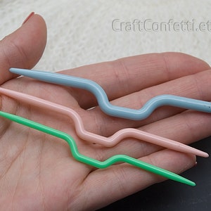 Puede incluir: Tres ganchillos de crochet de pl&aacute;stico en diferentes colores: azul claro, rosa y verde. Los ganchillos son curvos y tienen un extremo puntiagudo.