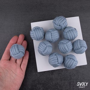 May include: Ten light blue decorative knots made from woven material. One knot is held in a hand, the others are on a white surface. The knots are evenly sized and have a textured appearance. The image includes the text "SVOLY CRAFT".