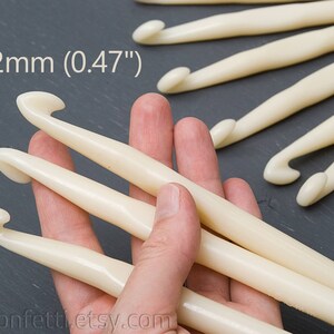 May include: A close-up of three ivory-colored crochet hooks, 12mm (0.47") in size, held in a hand. The hooks are made of smooth, polished plastic.
