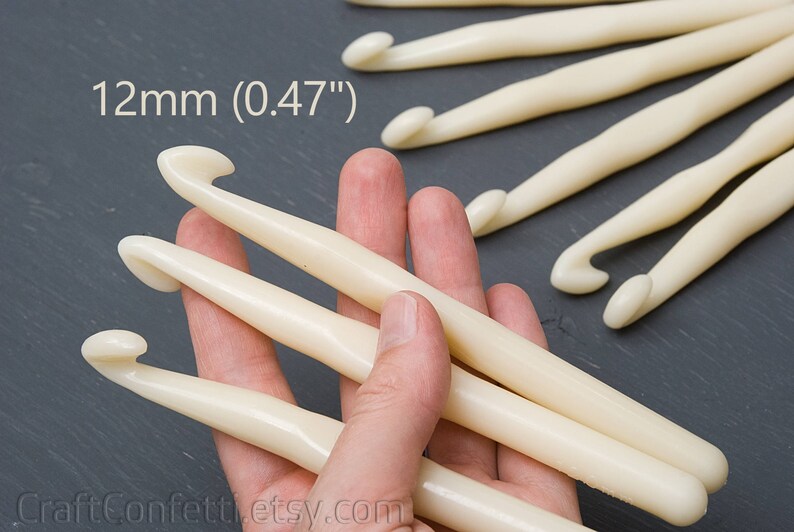 May include: A close-up of three ivory-colored crochet hooks, 12mm (0.47") in size, held in a hand. The hooks are made of smooth, polished plastic.