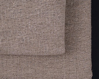 May include: Two pieces of tan burlap fabric, one folded over the other, against a black background. The fabric has a coarse, open weave, creating a textured surface. The neutral color and natural texture suggest a rustic or farmhouse aesthetic.