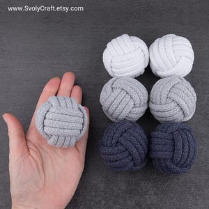 May include: Six spherical monkey fist knots made of woven rope, arranged in two columns. The colours include two white, two light grey, and two dark navy blue knots. One light grey knot is held in a palm to show the scale of the items.