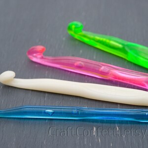 May include: Four crochet hooks in different colors: green, pink, white, and blue. The hooks are lying on a dark gray surface.