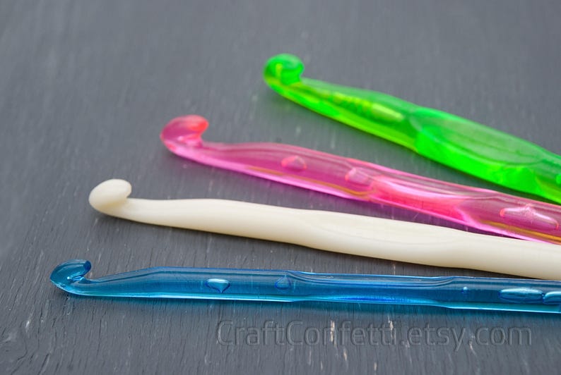May include: Four crochet hooks in different colors: green, pink, white, and blue. The hooks are lying on a dark gray surface.