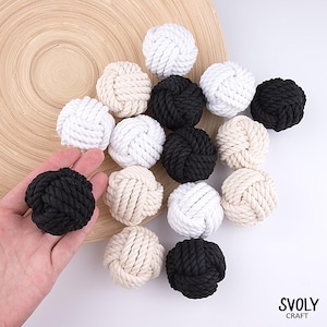 May include: A selection of decorative rope knots in white, black, and beige. The knots are displayed on a light wooden surface, with one black knot held in a hand. These nautical-themed knots are a craft item.