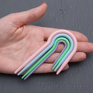 Puede incluir: Cuatro ganchillos de pl&aacute;stico de colores en rosa, azul claro, verde y morado dispuestos en arco&iacute;ris.