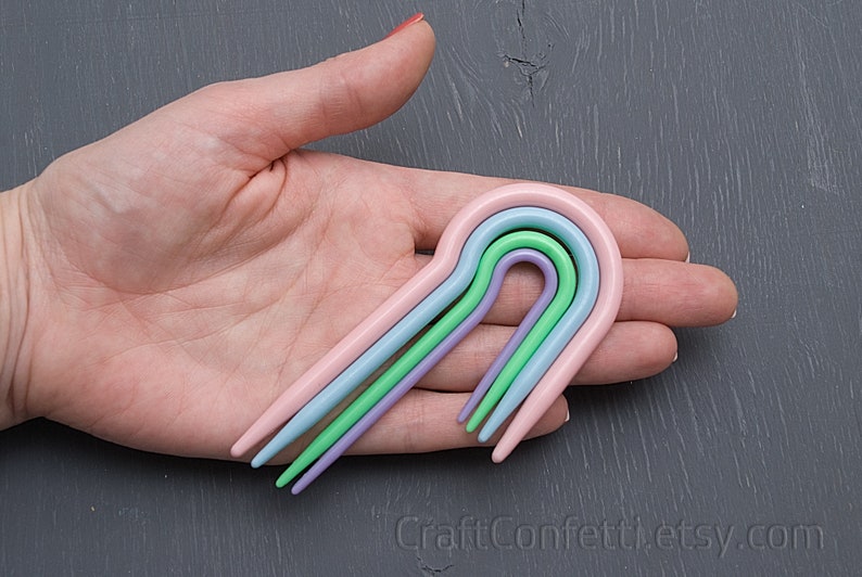Puede incluir: Cuatro ganchillos de pl&aacute;stico de colores en rosa, azul claro, verde y morado dispuestos en arco&iacute;ris.