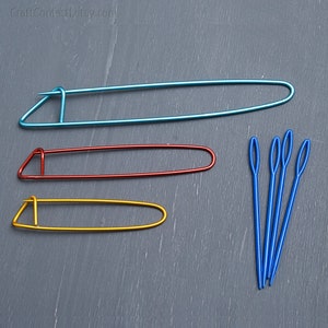 May include: A collection of knitting tools, including three colorful stitch holders in blue, red, and yellow, along with several blue yarn needles. The tools are arranged on a gray surface. The Etsy shop name "CraftConfetti.etsy.com" is visible in the top left corner.