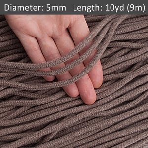 Puede incluir: Primer plano de un cordón de algodón marrón, de aproximadamente 5 mm de diámetro, con una mano sujetando algunos hilos. El cordón tiene 10 yardas (9 metros) de largo.