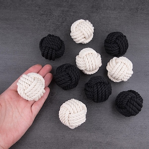 Può includere: Una collezione di nodi decorativi in corda nera e bianco sporco. Un nodo è tenuto in una mano. I nodi sono disposti su una superficie grigio scuro. I nodi sono fatti di corda spessa e intrecciata.