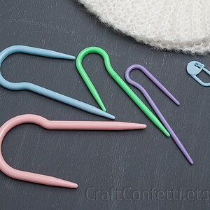Puede incluir: Cinco marcadores de punto de tejido de pl&aacute;stico de colores en varias formas, incluyendo una forma de gancho y una forma de c&iacute;rculo. Los marcadores est&aacute;n sobre una superficie gris oscuro. CraftConfettiEtsy.com est&aacute; impreso en la superficie.