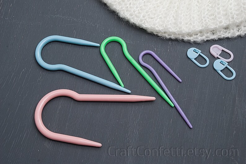 Puede incluir: Cinco marcadores de punto de tejido de pl&aacute;stico de colores en varias formas, incluyendo una forma de gancho y una forma de c&iacute;rculo. Los marcadores est&aacute;n sobre una superficie gris oscuro. CraftConfettiEtsy.com est&aacute; impreso en la superficie.