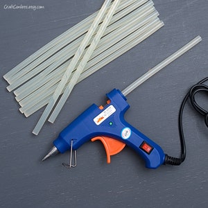 Puede incluir: Una pistola de pegamento caliente azul y naranja con un pegamento transparente insertado. La pistola de pegamento está enchufada y tiene un interruptor de encendido. Hay varios pegamentos transparentes apilados junto a la pistola de pegamento.