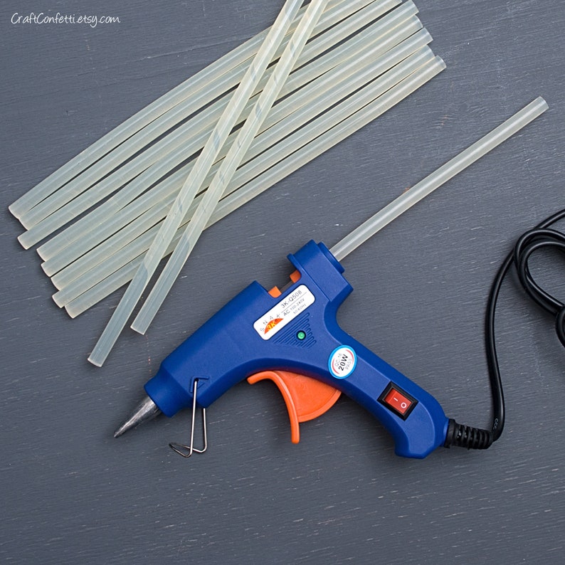 May include: A blue and orange hot glue gun with a clear glue stick inserted. The glue gun is plugged in and has a power switch. There are multiple clear glue sticks stacked next to the glue gun.