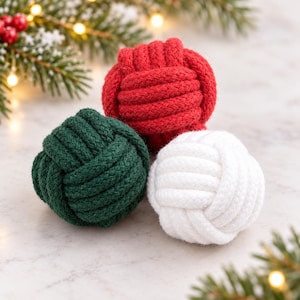 May include: Three spherical monkey fist knots made of thick woven rope in solid red, dark green, and white colours. The knots are arranged in a cluster, showcasing the intricate texture of the braided cord construction.
