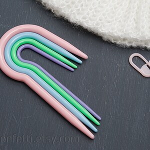 Puede incluir: Conjunto de cuatro ganchillos de pl&aacute;stico de colores vivos en rosa, azul, verde y morado. Los ganchillos est&aacute;n dispuestos en forma de arco&iacute;ris y est&aacute;n sobre una superficie gris oscuro.