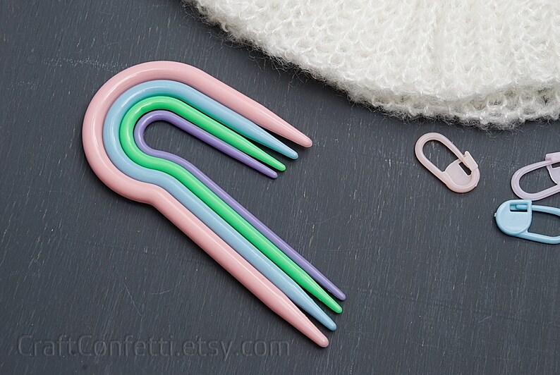 Puede incluir: Conjunto de cuatro ganchillos de pl&aacute;stico de colores vivos en rosa, azul, verde y morado. Los ganchillos est&aacute;n dispuestos en forma de arco&iacute;ris y est&aacute;n sobre una superficie gris oscuro.