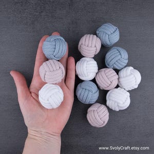May include: A selection of decorative knot balls in shades of light blue, pink, and white. The balls are crafted from a woven material and are displayed on a grey surface. The Etsy shop URL is visible in the bottom right corner.