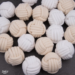 May include: A collection of decorative rope knot balls in cream and white. The balls are tightly knotted, creating a textured surface. The SVOLY CRAFT logo is visible in the bottom left corner. These could be used for home decor or craft projects.