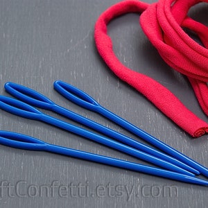Puede incluir: Cuatro agujas de lana de pl&aacute;stico azul sobre una superficie gris junto a un rollo de tela roja.