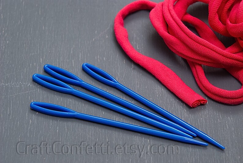 Puede incluir: Cuatro agujas de lana de pl&aacute;stico azul sobre una superficie gris junto a un rollo de tela roja.