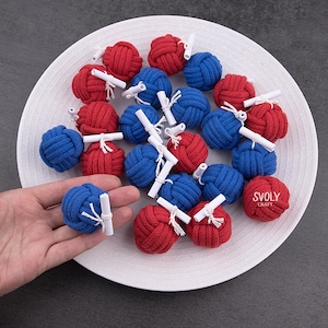 Könnte beinhalten: Eine Ansammlung roter und blauer dekorativer Knoten, jeweils mit einer kleinen weißen Schriftrolle versehen, angeordnet auf einem weißen Teller. Die Knoten bestehen aus dicht gewebtem Seil. Der Text "SVOLY CRAFT" ist enthalten.