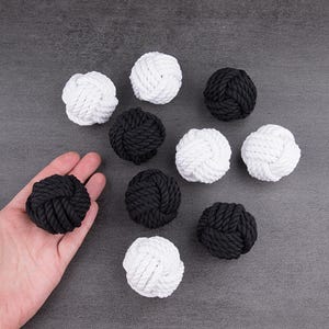 May include: A collection of ten spherical monkey fist knots made from twisted rope, featuring five solid white knots and five solid black knots. Each knot is constructed from a single continuous length of rope wound into a tight, textured ball.