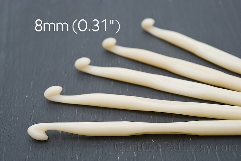 May include: Five ivory crochet hooks, each with a different length, laid out on a dark gray surface. The hooks are 8mm (0.31") in size.