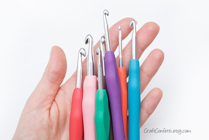 May include: A set of six colorful crochet hooks with metal tips. The hooks are arranged in a hand, with the largest hook at the top and the smallest hook at the bottom.