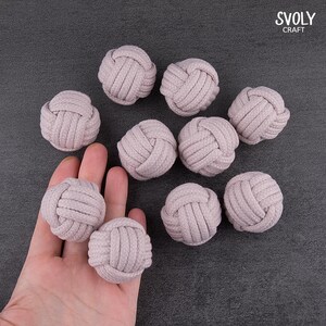 May include: A collection of ten light purple decorative rope knots. The knots are arranged on a dark gray surface, with some held in a hand. The knots are made of tightly woven rope.