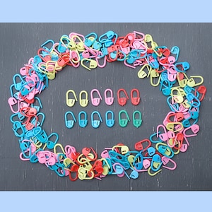 May include: A large assortment of colorful plastic stitch markers in various shapes and sizes. The markers are arranged in a circle around a smaller group of stitch markers in a variety of colors.
