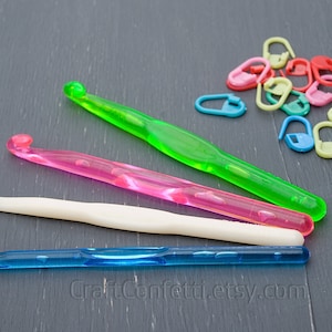 May include: Four colorful plastic crochet hooks and a pile of colorful plastic stitch markers on a gray wooden surface. The hooks are green, pink, white, and blue. The stitch markers are in various colors, including red, yellow, green, blue, and pink.