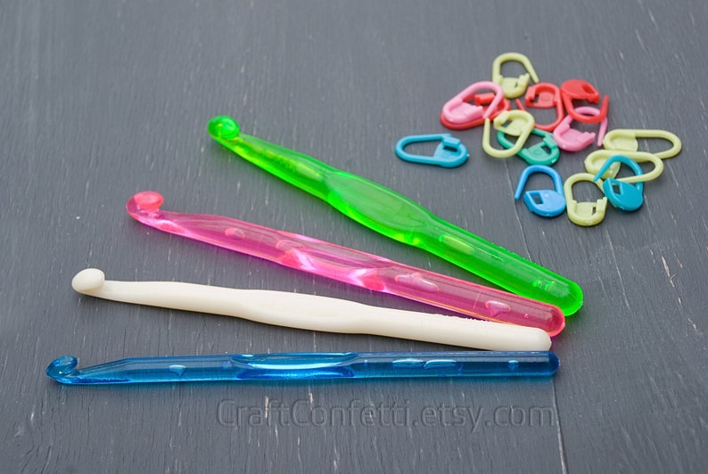 May include: Four colorful plastic crochet hooks and a pile of colorful plastic stitch markers on a gray wooden surface. The hooks are green, pink, white, and blue. The stitch markers are in various colors, including red, yellow, green, blue, and pink.