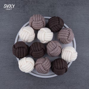 May include: A selection of decorative rope knots in shades of cream, brown, and grey, presented on a grey plate. The knots are intricately woven and appear handcrafted. The brand name "SVOLY CRAFT" is displayed in the top left corner.