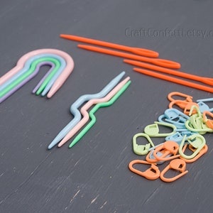 Puede incluir: Un conjunto de agujas de crochet de pl&aacute;stico de colores vivos y marcadores de punto. Las agujas de crochet son de varios tonos de azul, verde y rosa, mientras que los marcadores de punto son de tonos de verde, azul y naranja. Las agujas de crochet est&aacute;n dispuestas en forma de arco&iacute;ris, mientras que los marcadores de punto est&aacute;n en una pila.