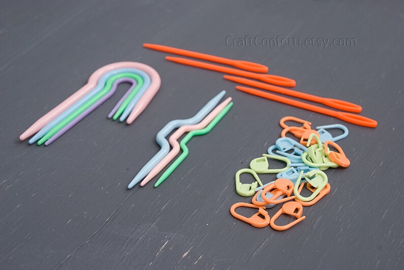 Puede incluir: Un conjunto de agujas de crochet de pl&aacute;stico de colores vivos y marcadores de punto. Las agujas de crochet son de varios tonos de azul, verde y rosa, mientras que los marcadores de punto son de tonos de verde, azul y naranja. Las agujas de crochet est&aacute;n dispuestas en forma de arco&iacute;ris, mientras que los marcadores de punto est&aacute;n en una pila.