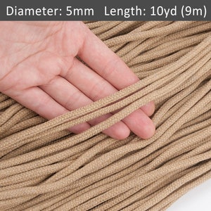 Puede incluir: Primer plano de un cordón de algodón beige de aproximadamente 5 mm de diámetro, con una mano sujetando una sección del cordón. El cordón tiene 10 yardas (9 metros) de largo.