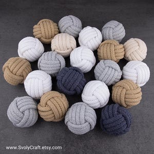 May include: A collection of decorative rope balls in various neutral colors, including white, gray, and tan. The balls are tightly knotted, creating a textured surface. The arrangement is on a dark gray background. The website address www.SvolyCraft.etsy.com is visible.